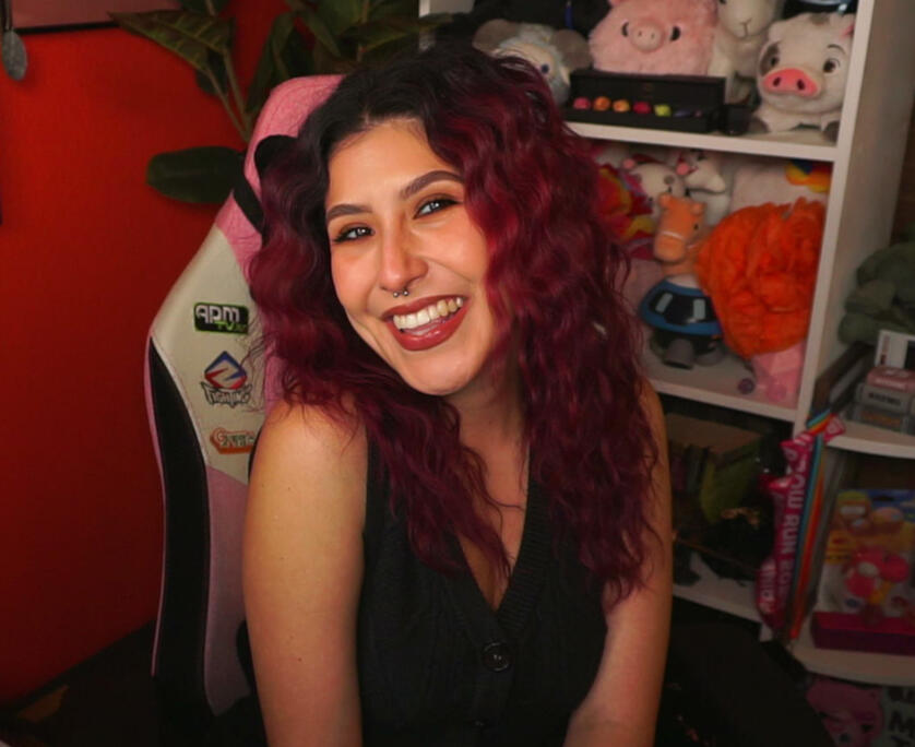 Bailee, a white Latina with deep red hair that is crimped. She is smiling at the camera and wearing a black knit vest.